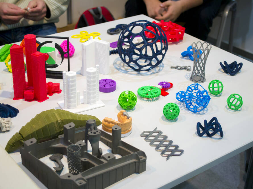 Ant stalo išdėstyti įvairūs spalvoti 3D spausdinti objektai - geometrinės figūros, pavaros, architektūriniai modeliai ir prototipai