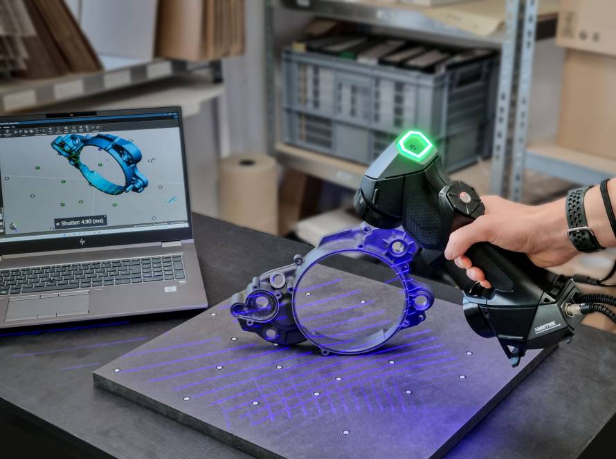 Rankinis 3D skeneris nuskaito mechaninę detalę, šalia atidarytas nešiojamas kompiuteris su modeliu ekrane