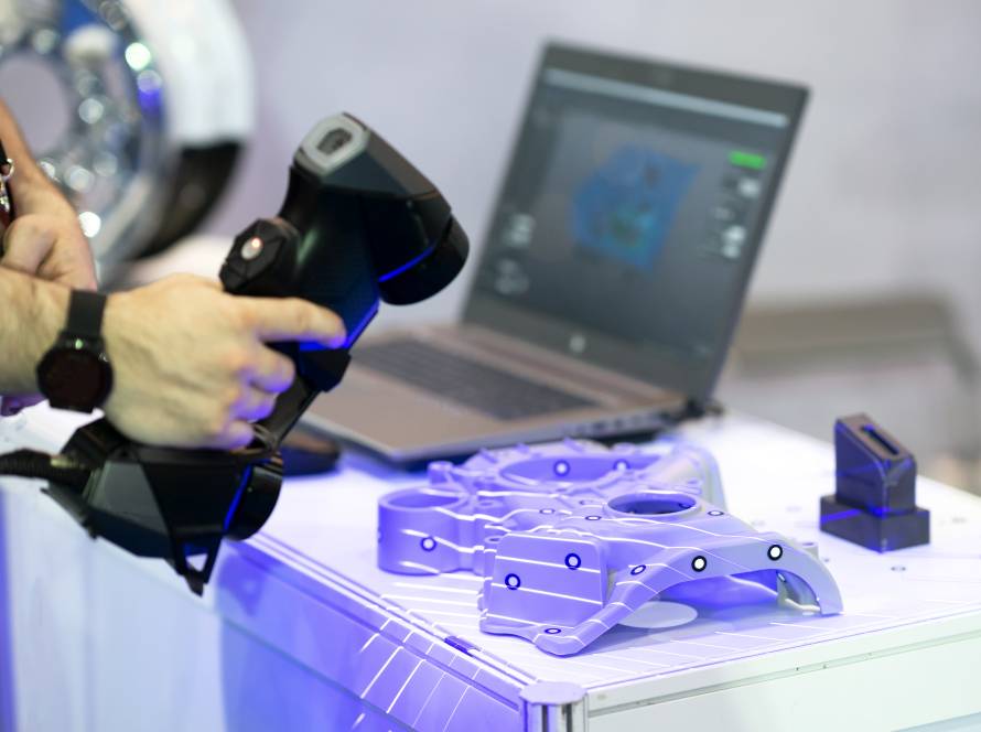Asmuo naudoja rankinį 3D skenerį mechaninei detalei skaitmeninti, šalia atidarytas nešiojamas kompiuteris su modelio peržiūra